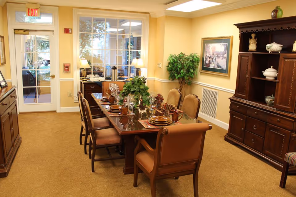 A warmly lit dining room with a long wooden table set for six people with plates, cups, and napkins. The room features beige walls, carpeted floor, a large wooden cabinet with decorative items, a framed painting on the wall, and a potted plant in the corner. There is a glass door and window at the far end letting in natural light.