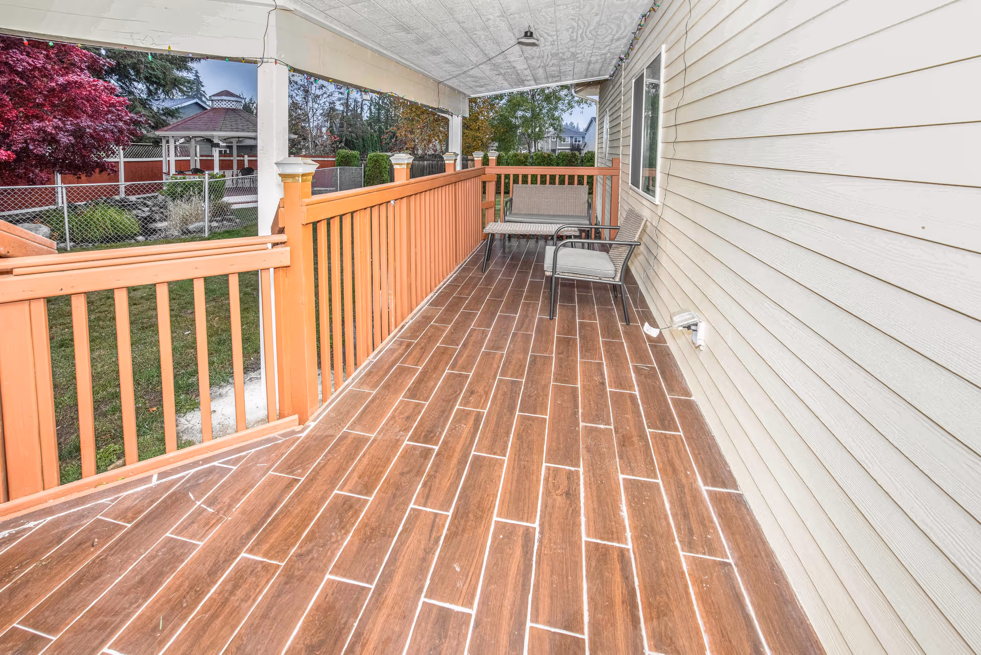 Covered porch with wood-look tile flooring, orange railing and outdoor seating overlooking a fenced yard and gazebo.