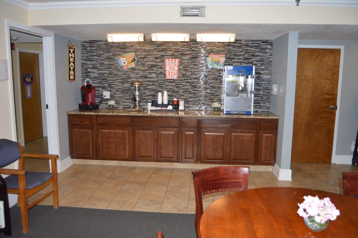 A kitchenette area with a tiled backsplash featuring a popcorn and movie theme. There is a popcorn machine, a coffee maker, a water dispenser, and cups on the granite countertop. Wooden cabinets are below the countertop. To the left is a wooden chair with blue cushions, and in the foreground is a round wooden table with a small vase of pink flowers. The walls are painted gray and beige, and there are two wooden doors visible.