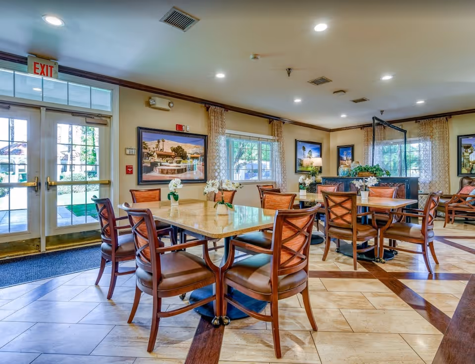 Interior view of a dining area in a senior living facility with multiple wooden tables and chairs arranged neatly. The room has large windows with patterned curtains, framed pictures on the walls, and a glass door with an exit sign above it. The floor is tiled with a decorative pattern, and there are small flower arrangements on the tables.
