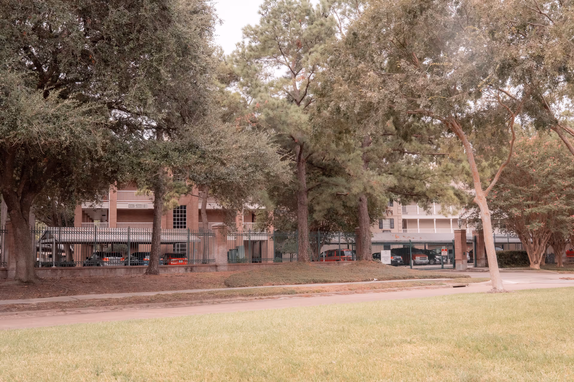 View of a residential building partially obscured by tall trees and a black metal fence. Several parked cars are visible behind the fence, and there is a grassy area and sidewalk in the foreground.