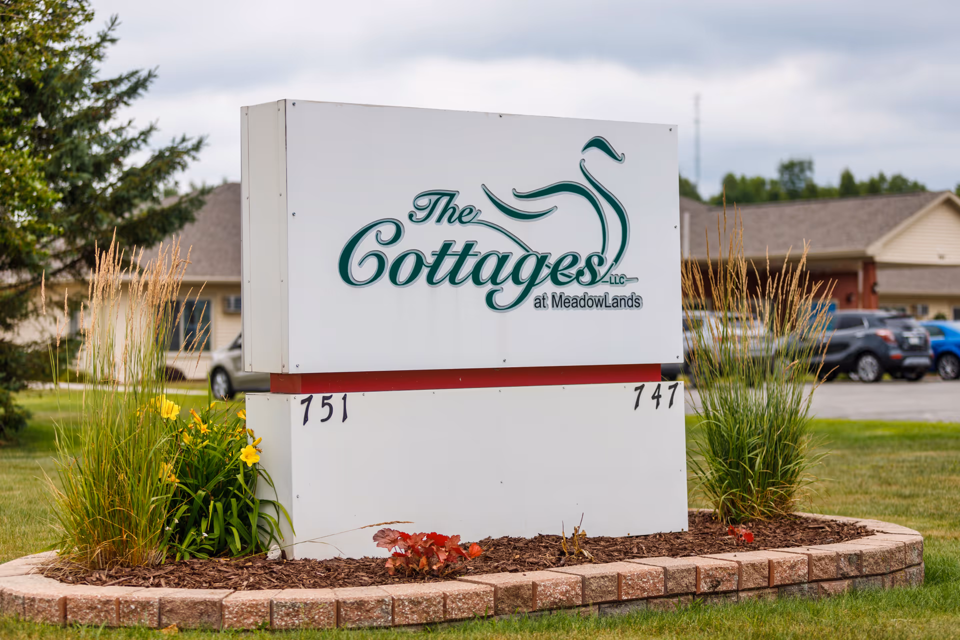 A large white sign with green text reading 'The Cottages LLC at MeadowLands' is displayed outdoors on a landscaped area with grass, plants, and flowers. In the background, there are residential buildings and parked cars under a cloudy sky.