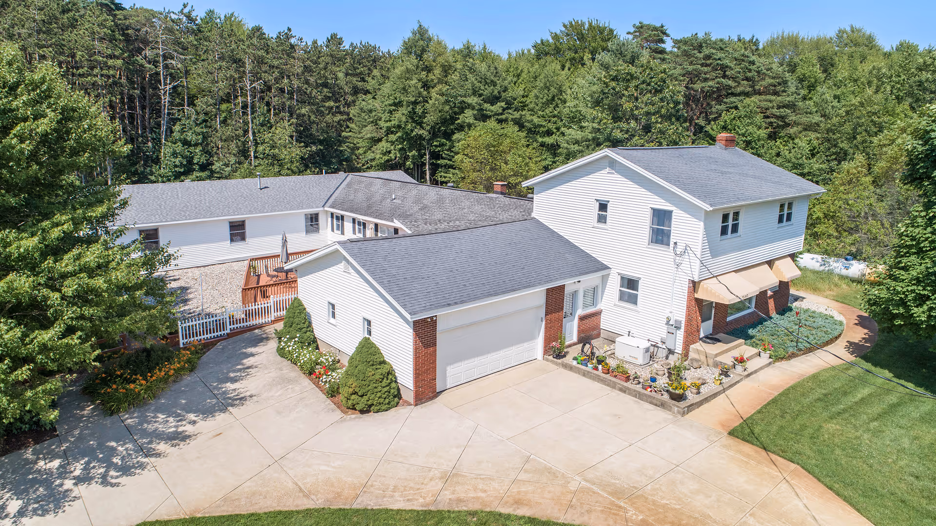 Aerial view of a two-story white house with a gray roof surrounded by greenery and trees. The house has a driveway and a small garden area with flowers and shrubs. There is a detached garage connected to the house and a paved walkway leading to the entrance.