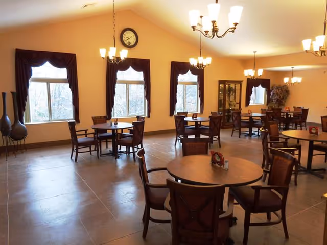 A spacious dining room with several round wooden tables and chairs arranged neatly. The room has large windows with dark curtains, beige walls, and multiple chandeliers providing warm lighting. There is a clock on the wall and a glass cabinet in the corner.