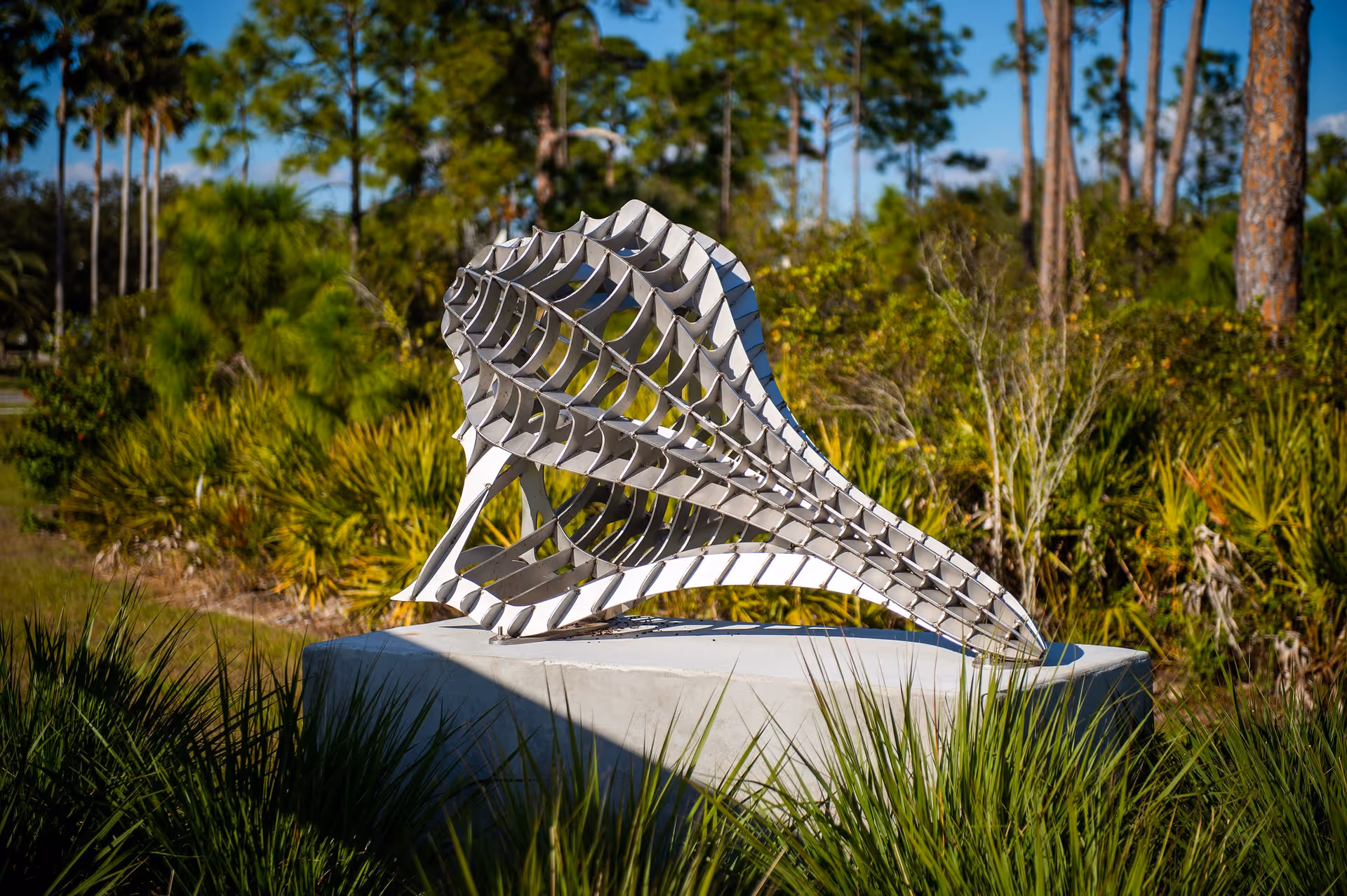 A modern abstract metal sculpture resembling a curved, open lattice structure is displayed outdoors on a concrete pedestal surrounded by green grass and trees under a clear blue sky.
