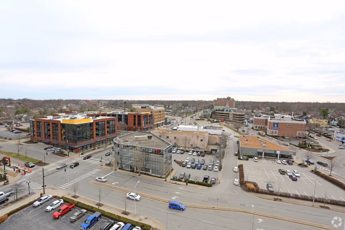 Aerial view of a commercial area with multiple buildings, parking lots, and streets. The scene includes a modern multi-story building with large windows, several smaller buildings, and numerous parked cars. The sky is overcast.