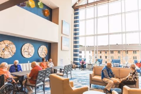 A bright and spacious common area in a senior living facility with large floor-to-ceiling windows letting in natural light. Several elderly residents are seated around tables and on sofas, engaging in conversation. The room features modern decor with blue accent walls, circular wall art, and patterned carpet.