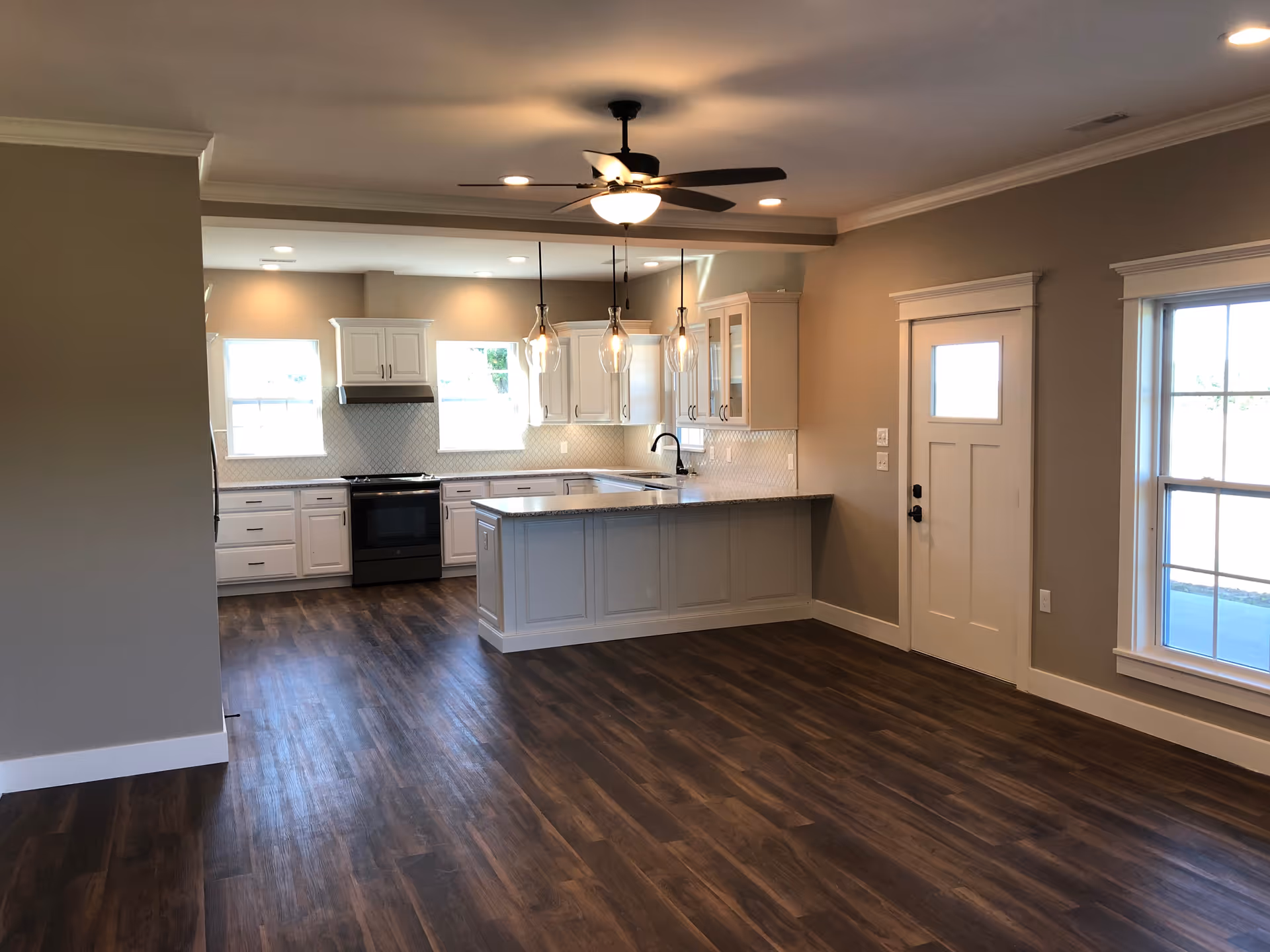 Interior view of a modern kitchen with white cabinets, a kitchen island with a granite countertop, three pendant lights hanging above the island, a ceiling fan with light, dark wood flooring, and beige walls. There are two windows above the kitchen counter and a white door with a window on the right side.