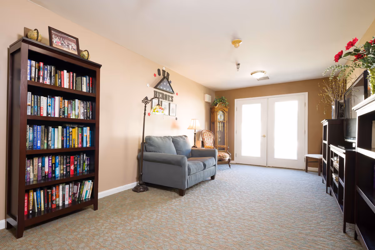 A cozy living room area with a blue loveseat, an antique wooden chair, a tall grandfather clock, and a dark wooden bookshelf filled with books. The room has beige walls, a patterned carpet, and double glass doors letting in natural light. There are decorative items on the bookshelf and a wall decoration that reads 'Our Heroes'.