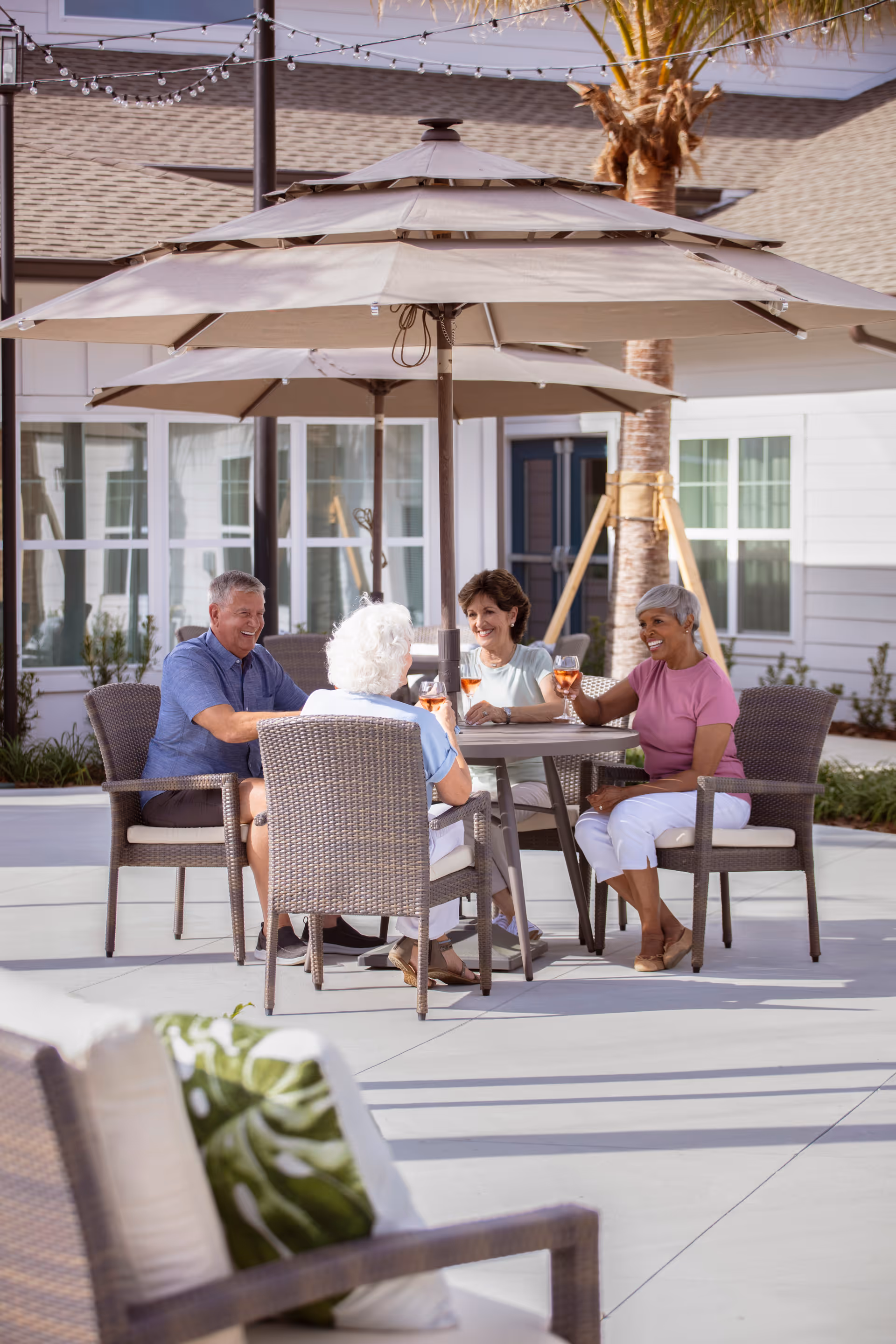 Four senior adults sitting around a patio table under a large umbrella, enjoying drinks and smiling in an outdoor courtyard area of a senior living facility.