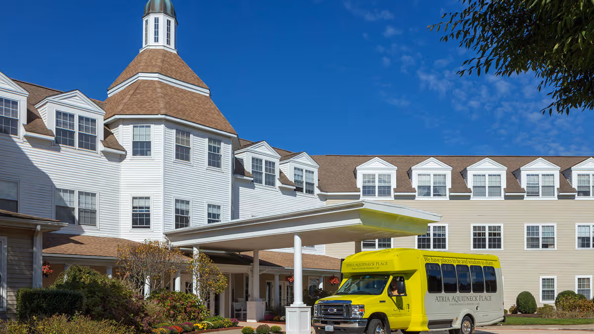 Exterior view of Atria Aquidneck Place senior living facility showing a large multi-story building with white and beige siding, multiple windows, and a distinctive turret. A covered entrance is visible with a yellow shuttle bus parked underneath. The sky is clear and blue.