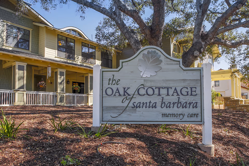 Exterior view of Oak Cottage of Santa Barbara Memory Care facility with a large wooden sign in the foreground displaying the facility's name. The building behind has multiple windows, a porch with hanging plants, and is surrounded by trees and landscaping.