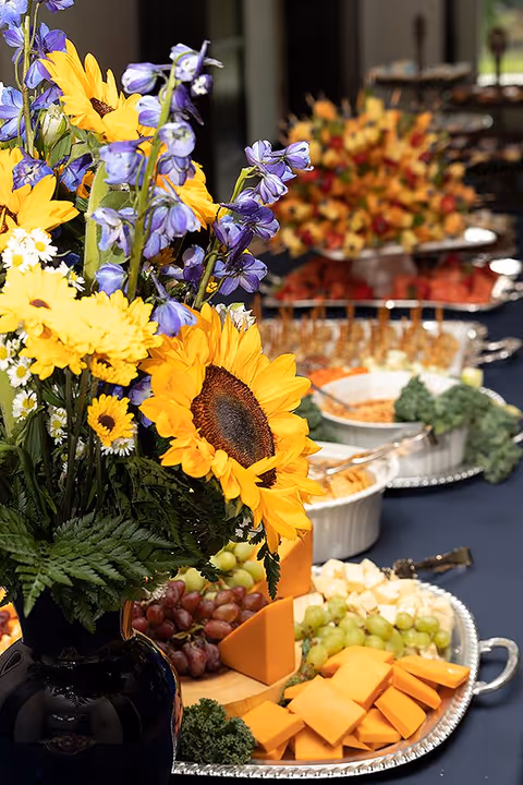A close-up view of a floral arrangement with sunflowers, yellow daisies, and purple flowers in a black vase, placed on a table with a variety of food platters including cheese, grapes, and other appetizers in the background.