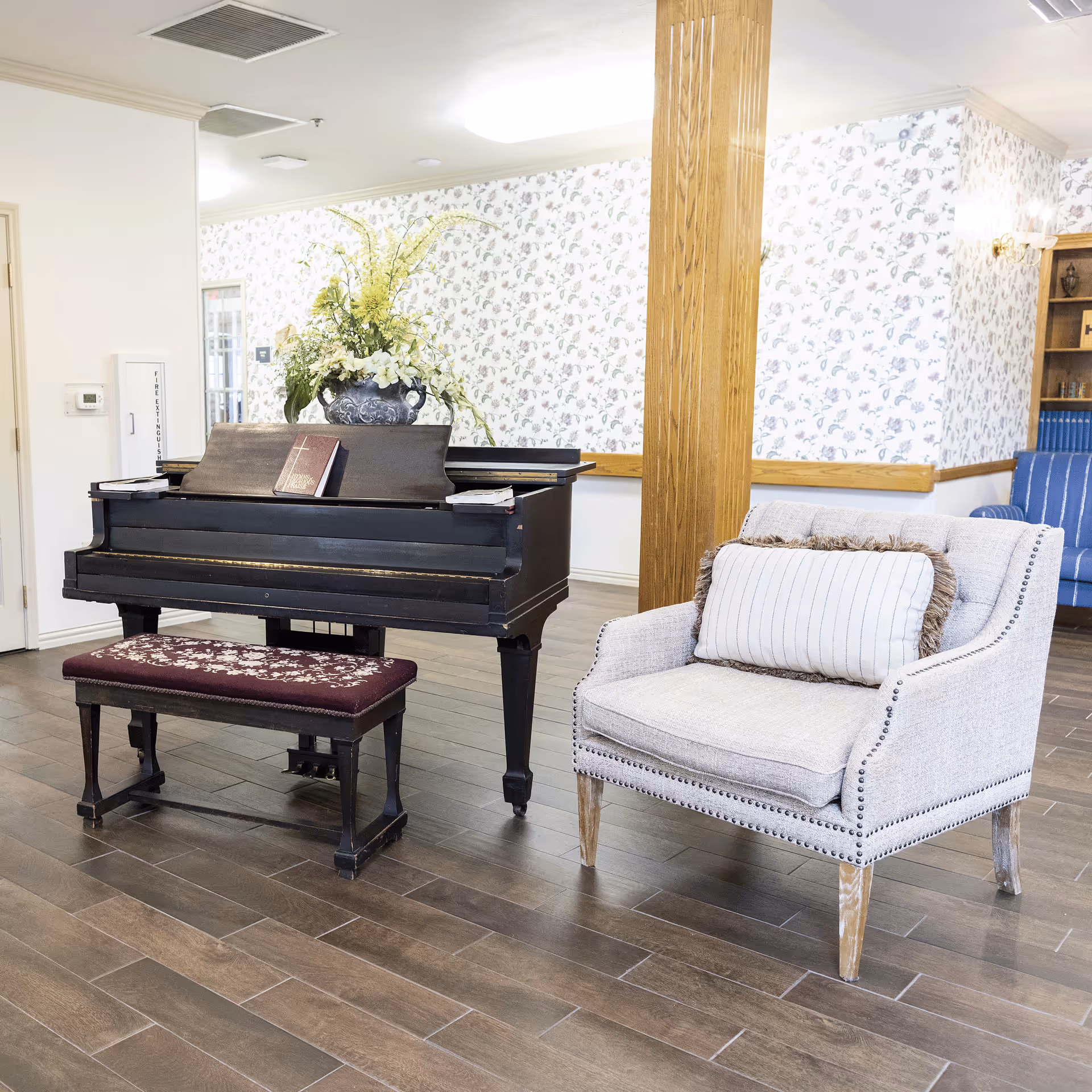 Interior of a senior living facility featuring a black upright piano with a floral arrangement on top and a book resting on the music stand. Next to the piano is a beige upholstered armchair with a decorative pillow. The room has wood-look tile flooring, floral wallpaper, and a wooden column. In the background, there is a blue striped sofa and a wooden bookshelf.