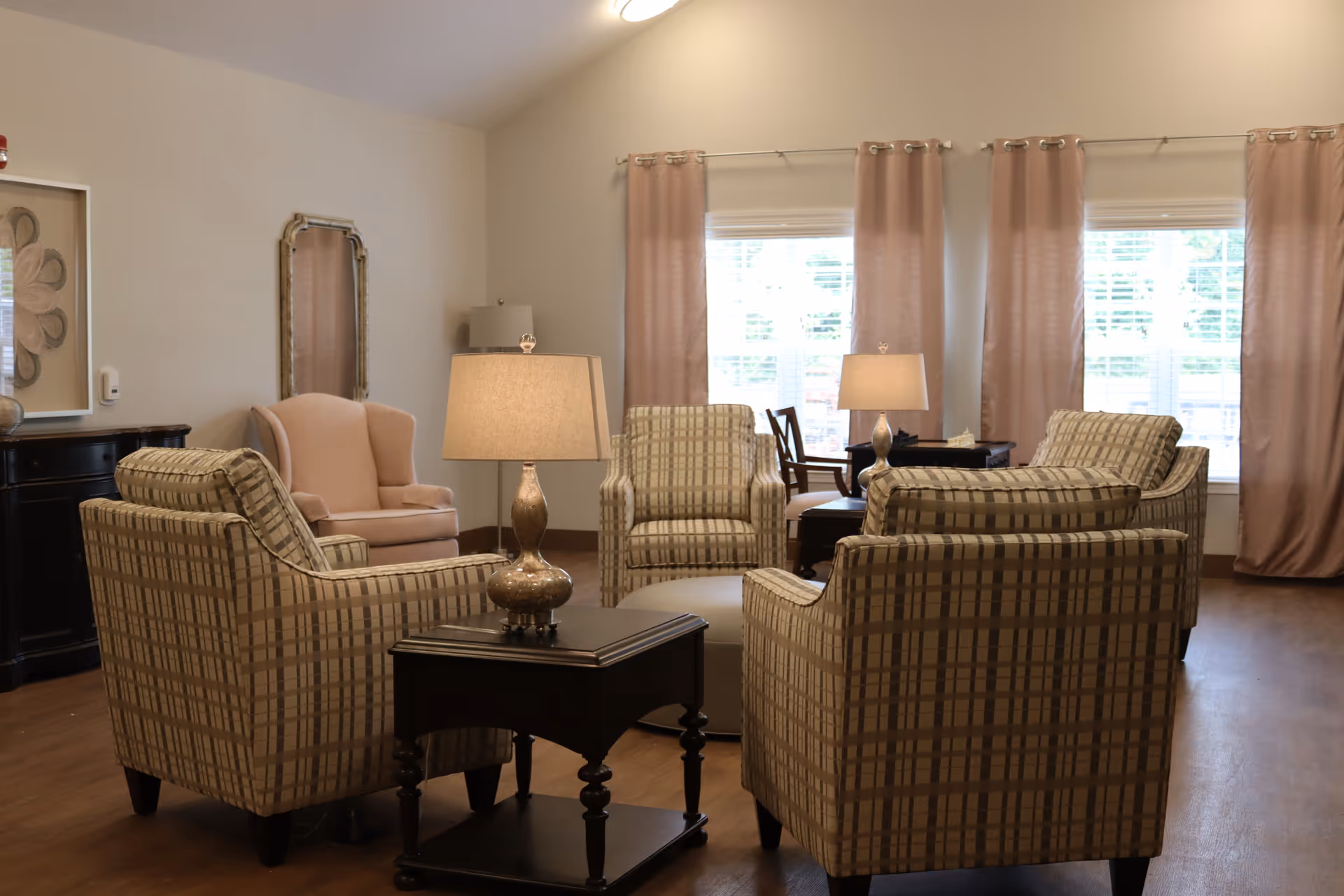 A cozy living room area with four plaid armchairs arranged around a round ottoman and two side tables with lamps. There is a pink upholstered armchair in the corner, a black sideboard with decorative items, a wall mirror, and large windows with pink curtains letting in natural light.