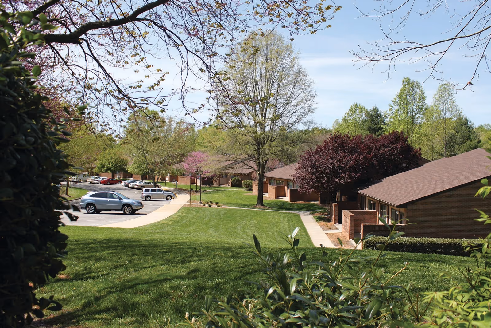 View of a senior living facility named Parkwood Place showing a well-maintained grassy lawn with trees and shrubs, a sidewalk, parked cars, and single-story brick buildings with brown roofs under a clear blue sky.