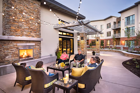 A group of people relax on wicker chairs around a table and outdoor fireplace in a lit courtyard of a multi-story senior living building.