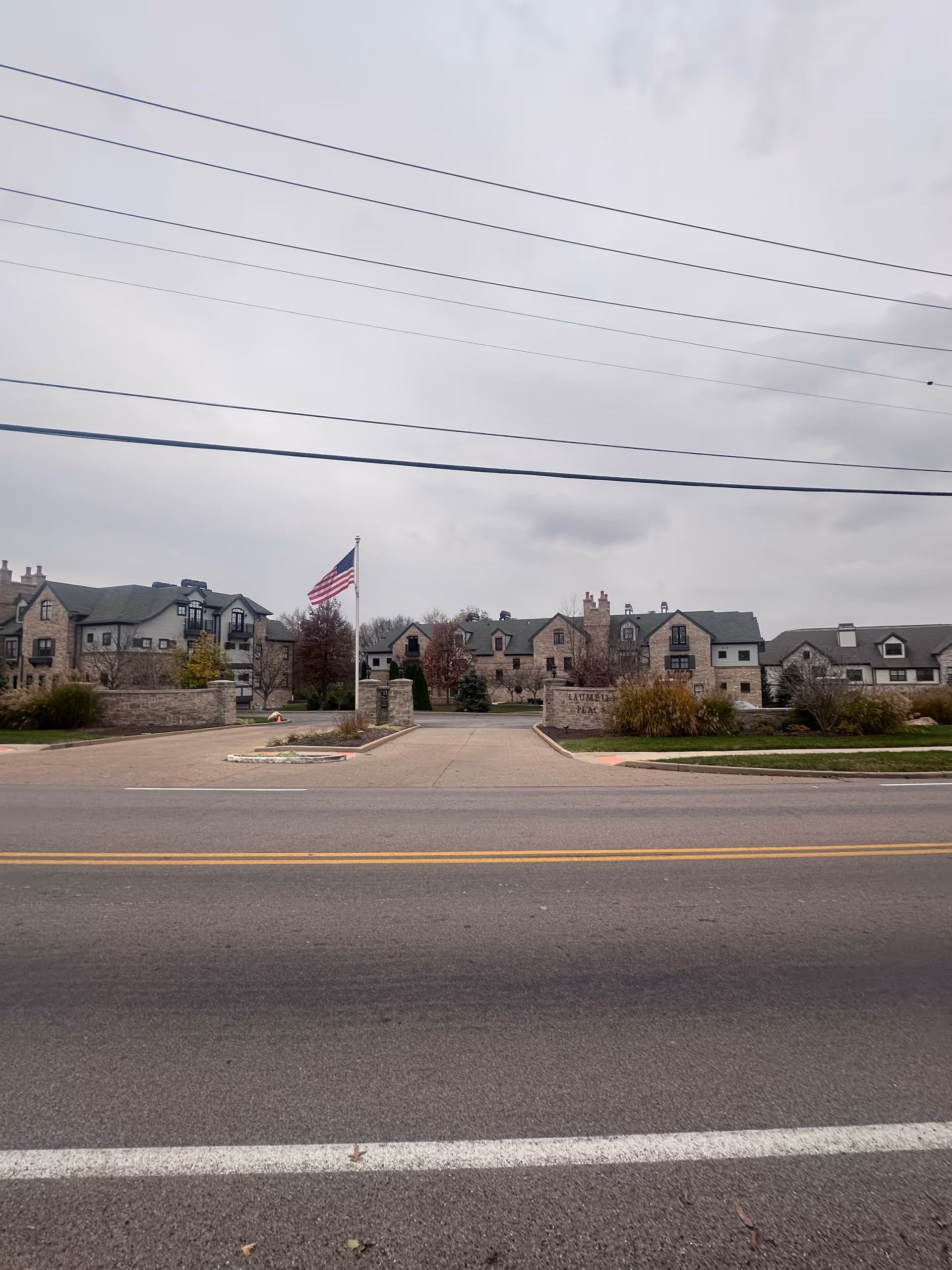 Stone-faced senior living complex entrance with an American flag and driveway seen from across the street under an overcast sky.