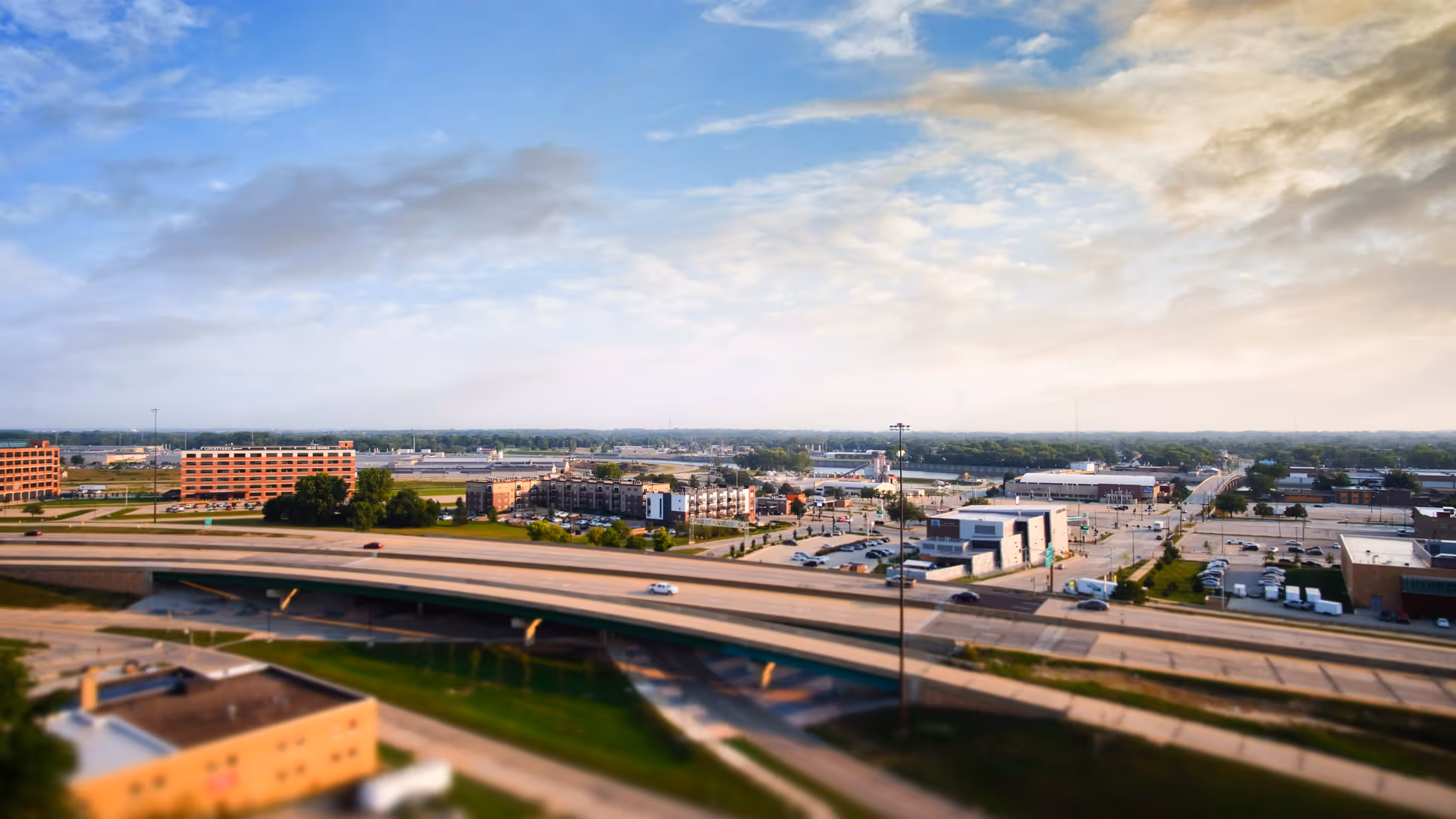 A wide aerial view of a cityscape featuring highways, buildings, parking lots, and a partly cloudy sky during daytime.
