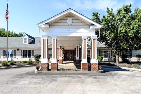Covered entrance/portico of a single-story senior living building with brick columns, a driveway, landscaping, and an American flag.