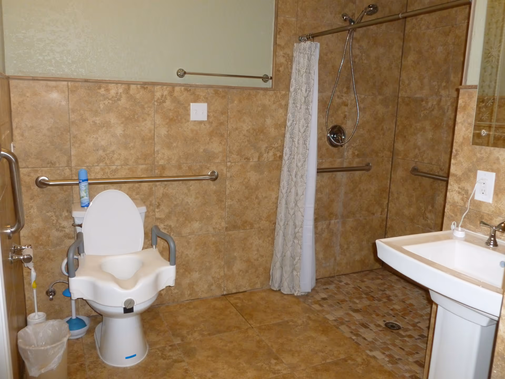 A bathroom with beige tiled walls and floor, featuring a toilet with arm supports and a raised seat, a white pedestal sink, a walk-in shower with a handheld showerhead and a patterned shower curtain, and multiple grab bars for accessibility.