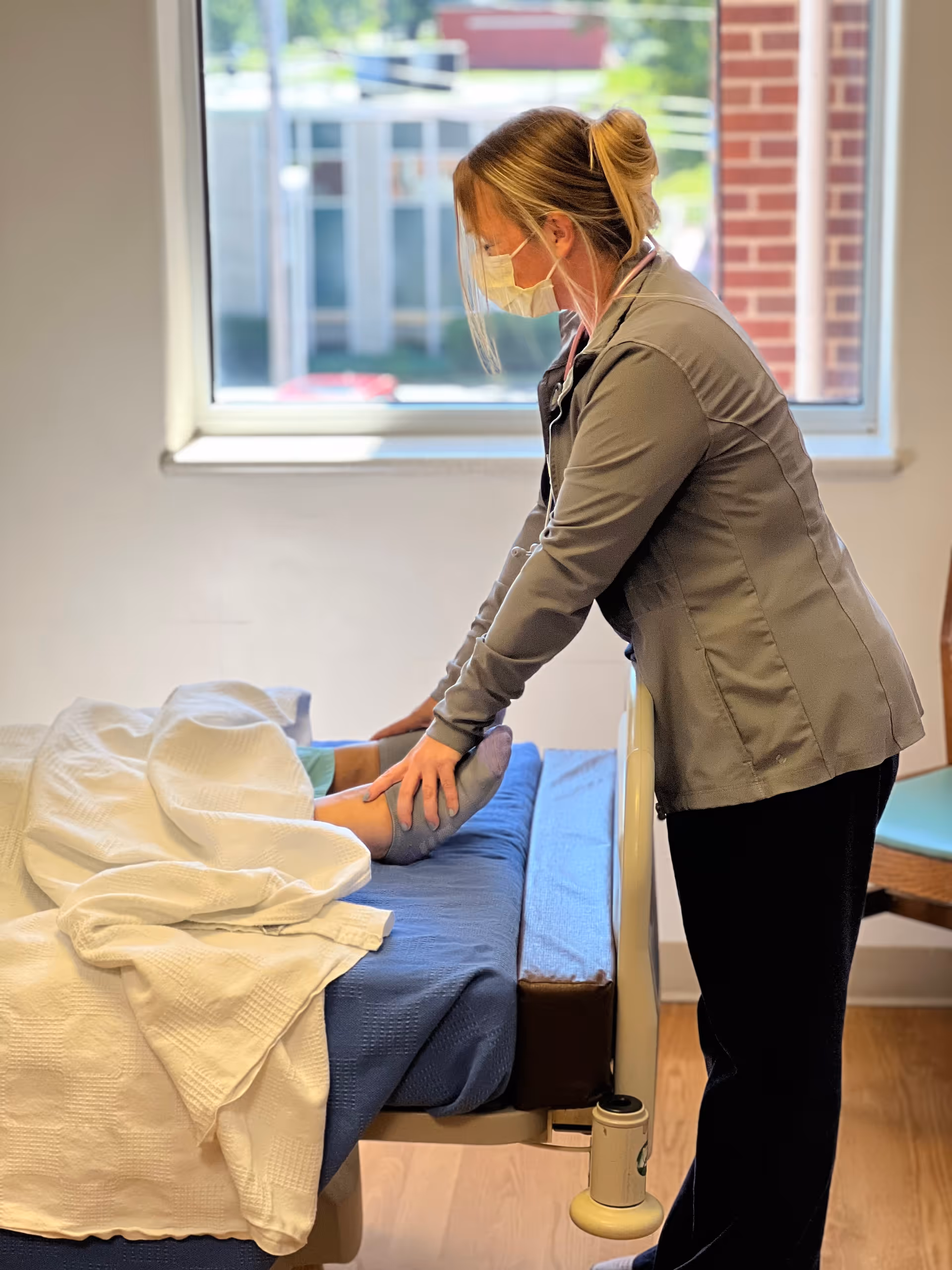 A healthcare worker wearing a mask and gray jacket is standing beside a hospital bed, gently holding the feet of a patient lying under a white blanket. The room has a window with a view of a building and some greenery outside.