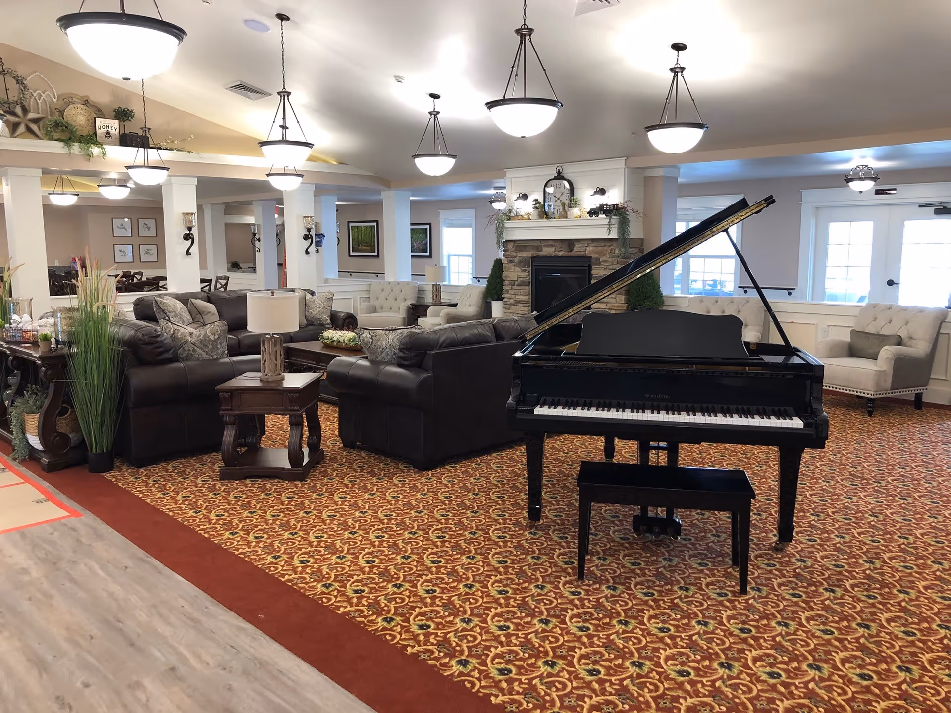 A spacious and well-lit living room area featuring a black grand piano with a matching bench in the foreground. The room has patterned carpet flooring and multiple seating arrangements including dark leather sofas, beige armchairs, and wooden side tables with lamps. A stone fireplace with decorative items and a clock is centered on the far wall. Several hanging ceiling lights illuminate the room, and large windows and glass doors allow natural light to enter.