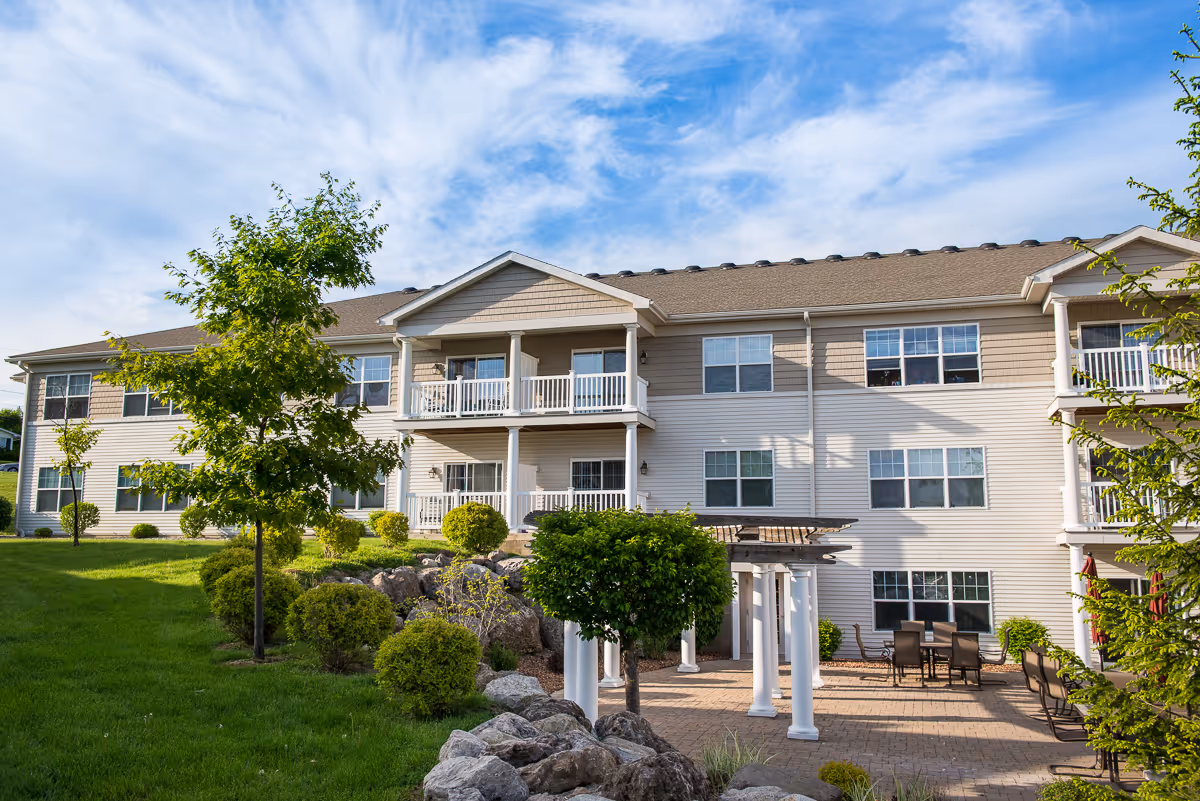 Exterior view of a two-story senior living facility building with beige siding and white trim. The building features balconies, multiple windows, and a patio area with outdoor seating and a pergola. The surrounding landscape includes green grass, trees, bushes, and rocks under a partly cloudy blue sky.