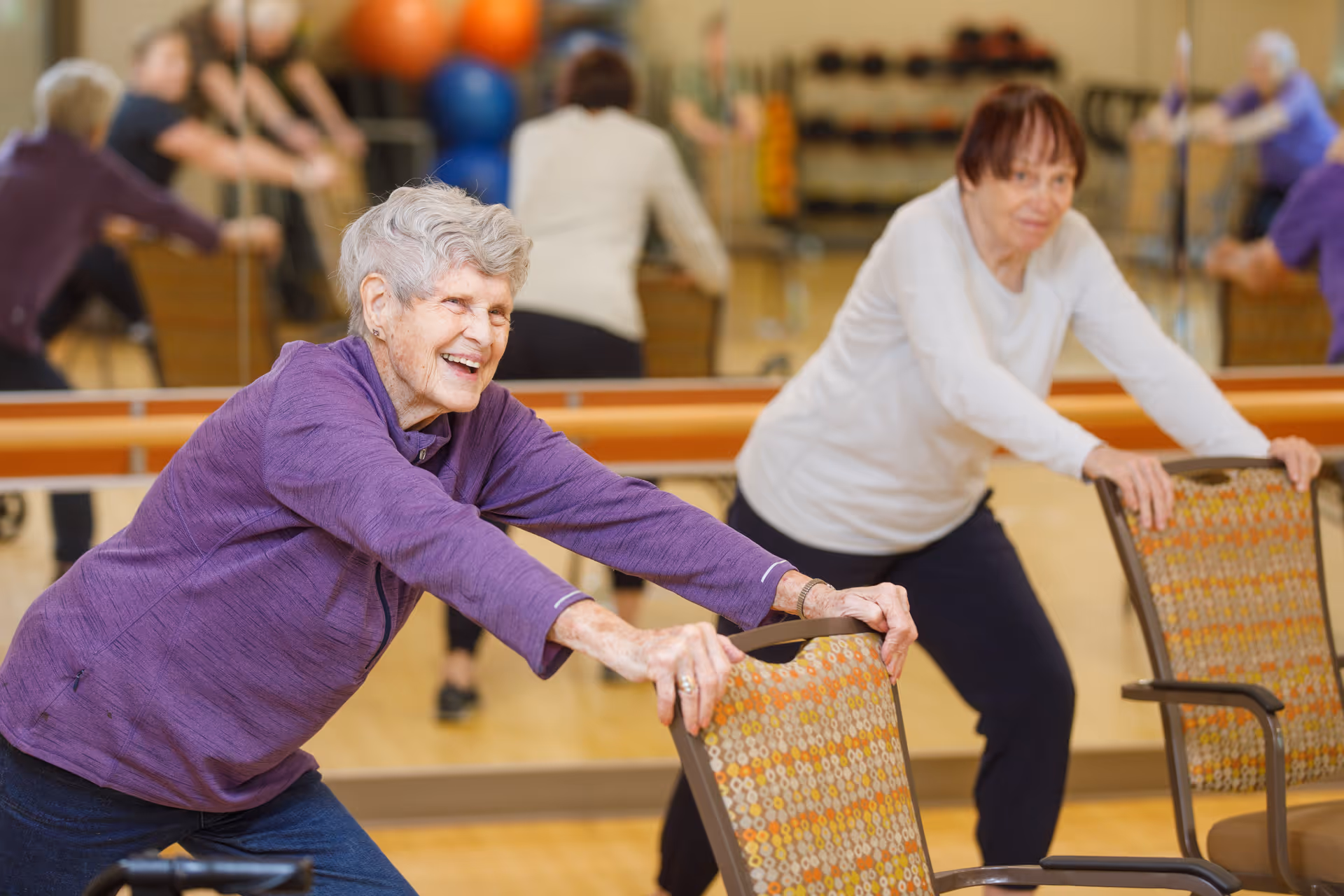 Two elderly women exercising in a fitness room, using chairs for support while stretching. One woman is smiling and wearing a purple jacket, and the other is wearing a white top. There are exercise balls and mirrors in the background.