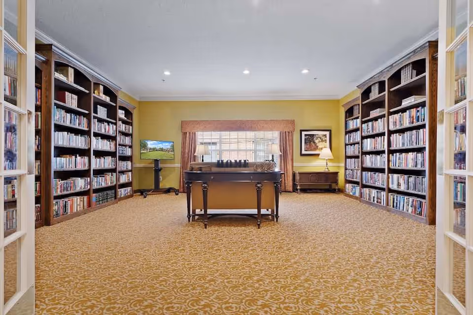 A spacious library room with yellow walls and patterned carpet. The room features tall wooden bookshelves filled with books on both sides. In the center, there is a wooden table with decorative items including a sign that spells 'HOME'. A window with curtains and two table lamps is at the back wall, along with a framed picture and a TV on a stand.