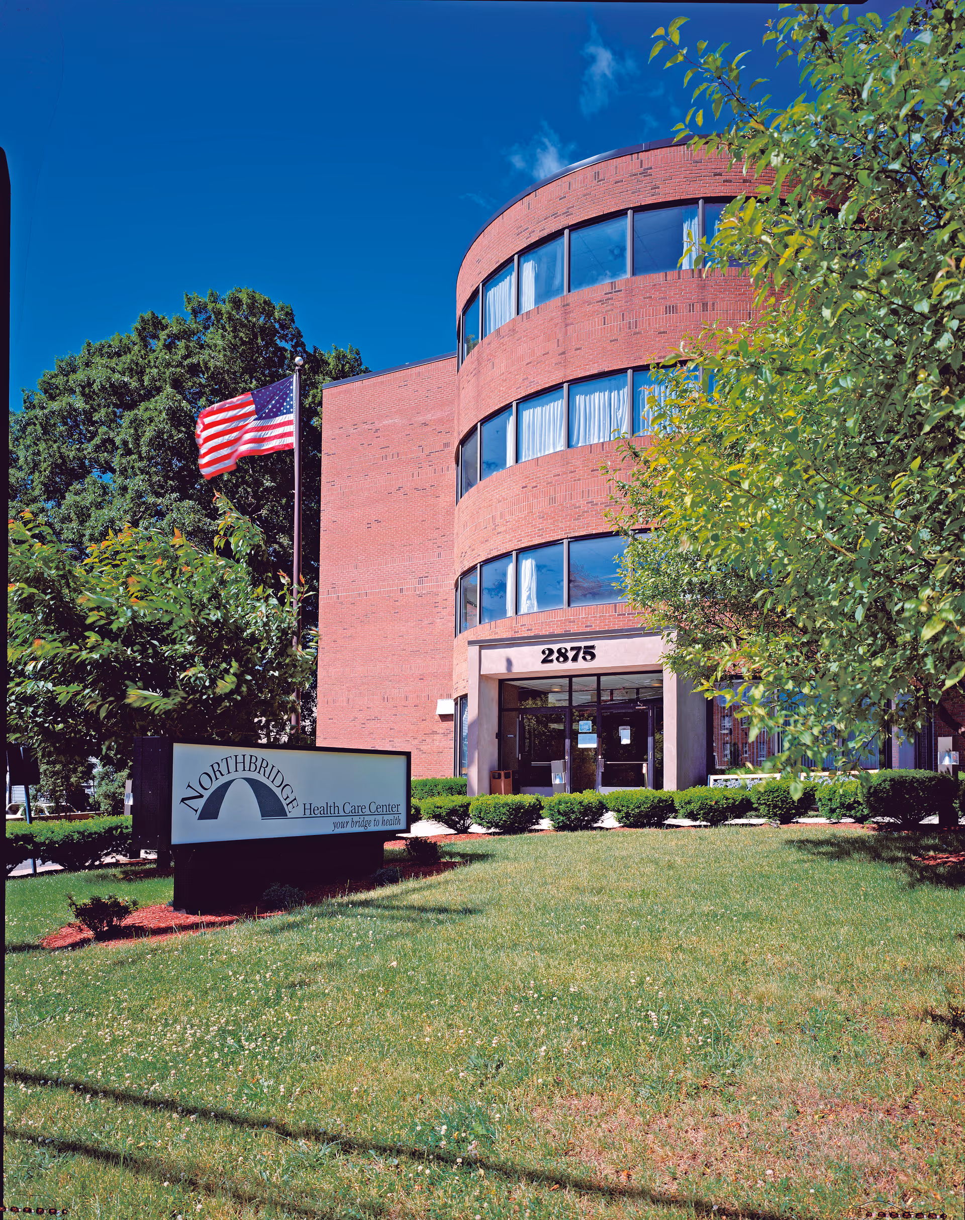 Brick three-story curved health care center building with an American flag, a lawn sign for Northbridge Health Care Center, and a grassy front lawn.