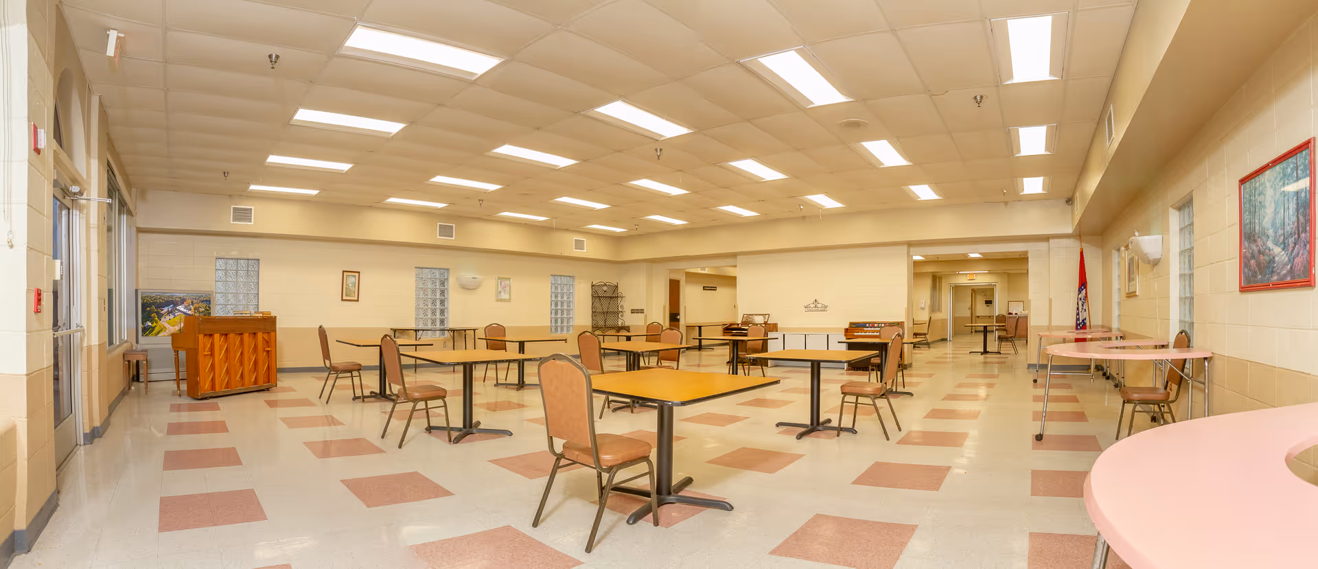 A spacious, well-lit room with multiple square tables and chairs arranged throughout. The floor has a checkered pattern with pink and white tiles. There is a piano against the left wall near a glass door, and several framed pictures hang on the walls. The ceiling has recessed lighting panels, and the room appears clean and organized, suitable for dining or group activities.