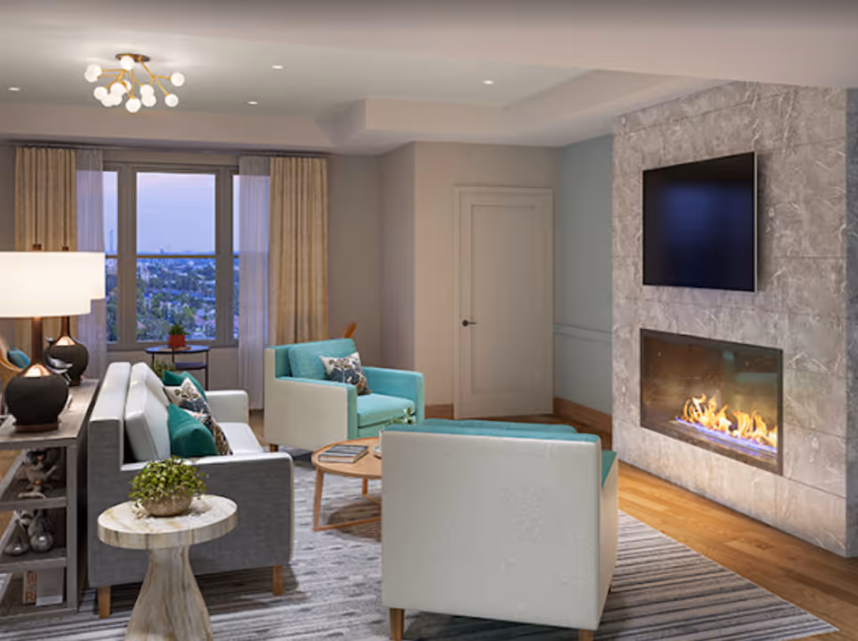 A modern living room with a gray sofa, two light blue armchairs, a round wooden coffee table, and a small round side table with a plant. There is a fireplace with a fire burning and a flat-screen TV mounted above it. Large windows with beige curtains offer a view of the city outside. The room has wooden flooring and a striped area rug.