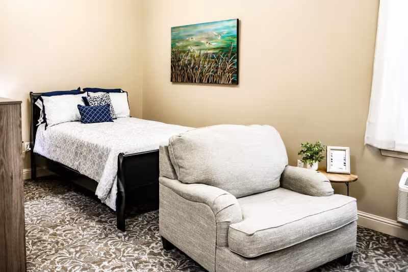 A furnished bedroom with a bed, upholstered armchair, small side table, and landscape wall art.