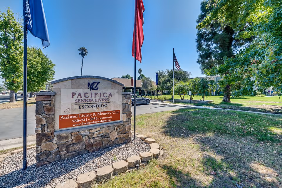 Outdoor view of the entrance sign for Pacifica Senior Living Escondido, surrounded by a stone base and three flagpoles with flags. The background shows a parking area, trees, and a clear blue sky.