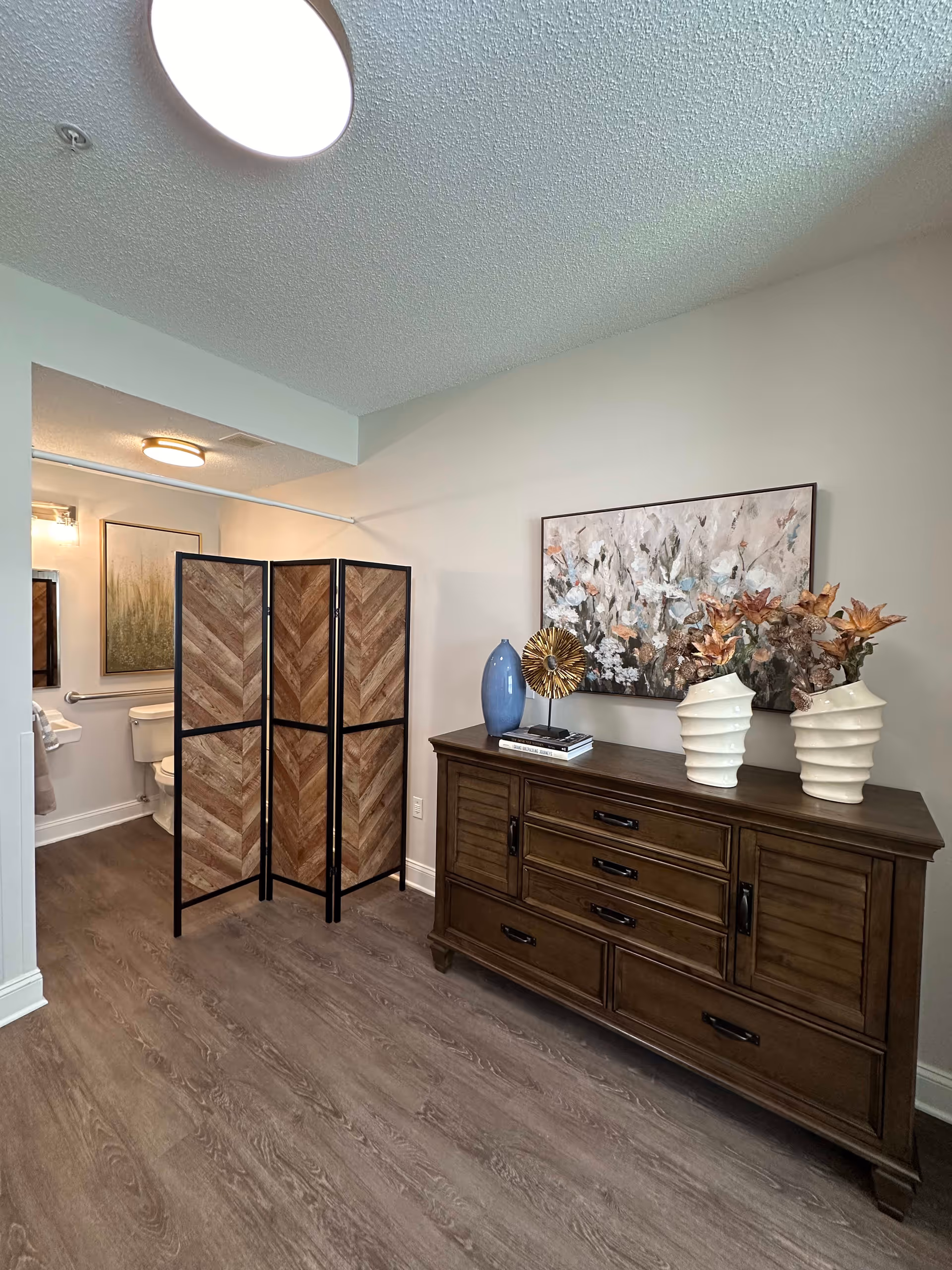 Interior view of a room with a wooden dresser topped with decorative items including two white vases with flowers, a blue vase, and a decorative gold object. A large floral painting hangs on the wall above the dresser. To the left, a wooden folding screen partially conceals a bathroom area with a toilet and framed artwork on the wall. The floor is wood-style laminate and the walls are painted light beige.