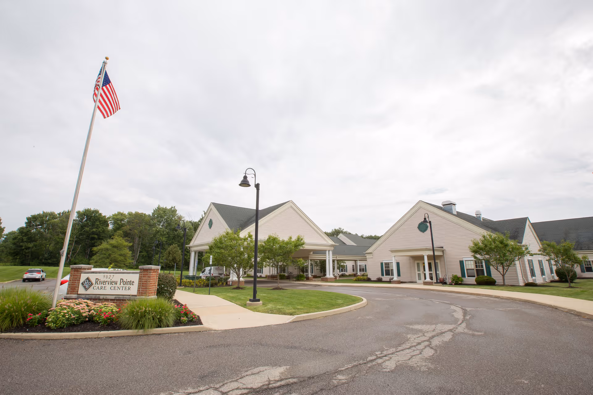Exterior view of Riverview Pointe Care Center showing a large building with beige siding and green shutters, a circular driveway, a flagpole with an American flag, and landscaped greenery under a cloudy sky.