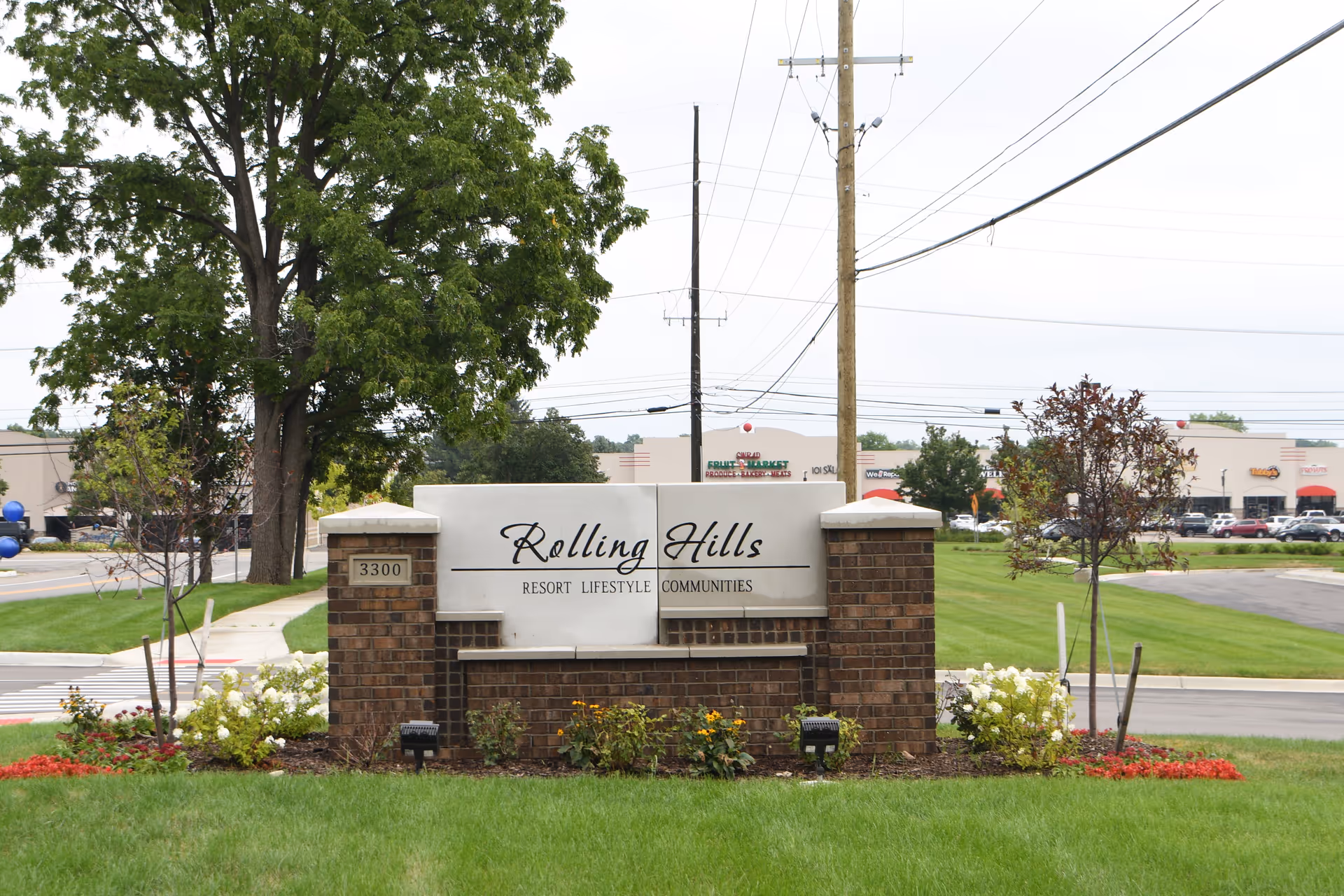 A brick and stone sign for Rolling Hills Resort Lifestyle Communities located on a grassy area with flowers and trees around it. In the background, there is a street and a shopping plaza with various stores and parked cars.