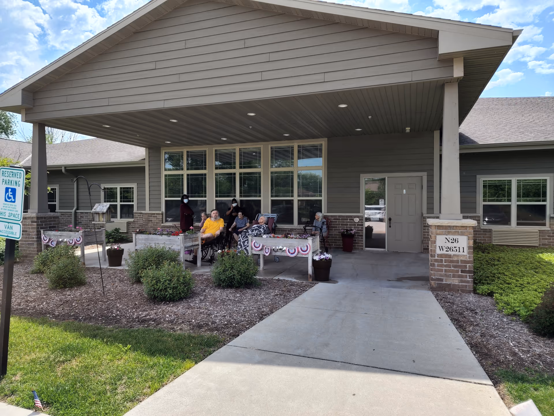 Entrance area of a senior living facility with a covered porch where several elderly people and a caregiver are seated and standing. The porch has raised garden beds decorated with patriotic bunting. There is a reserved parking sign for handicapped access visible on the left side, and the building exterior is gray with brick accents.