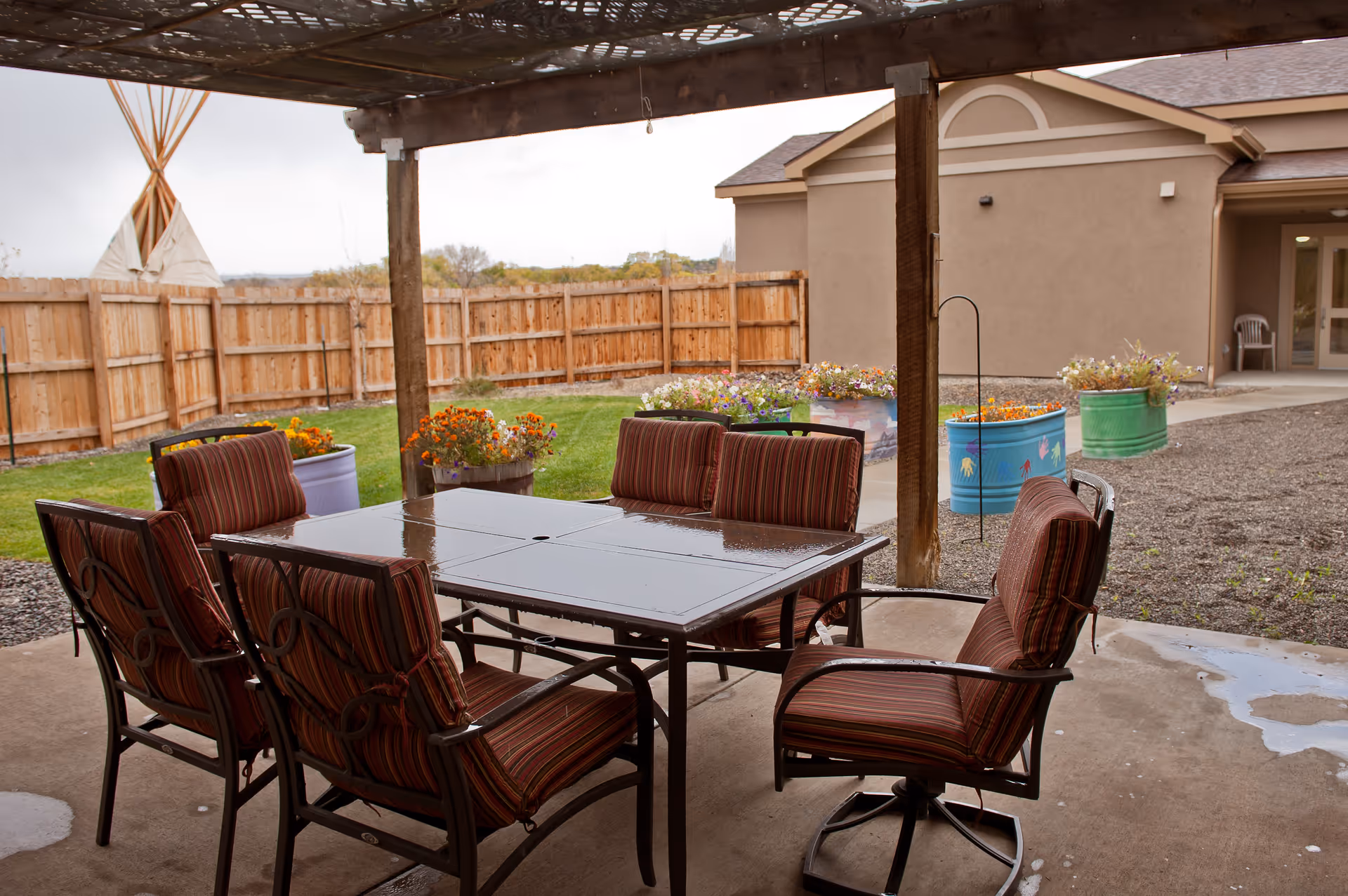 Outdoor covered patio area with a glass-top table and six cushioned chairs arranged around it. The patio overlooks a fenced yard with green grass, colorful flower pots, and a tipi in the corner. A beige building with an entrance is visible in the background.