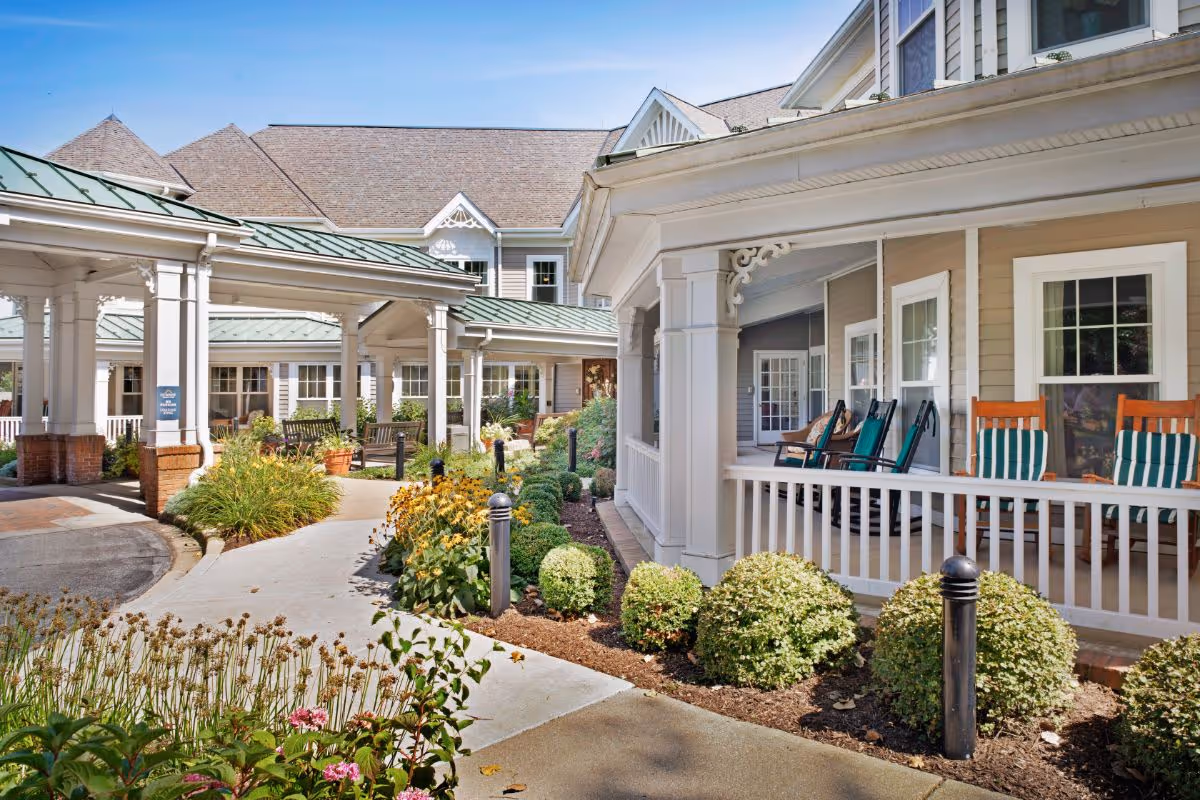 Exterior view of Sunrise of Flossmoor showing a covered walkway with white columns and green metal roofing, a garden with bushes and flowers, and a porch with rocking chairs and striped cushions.
