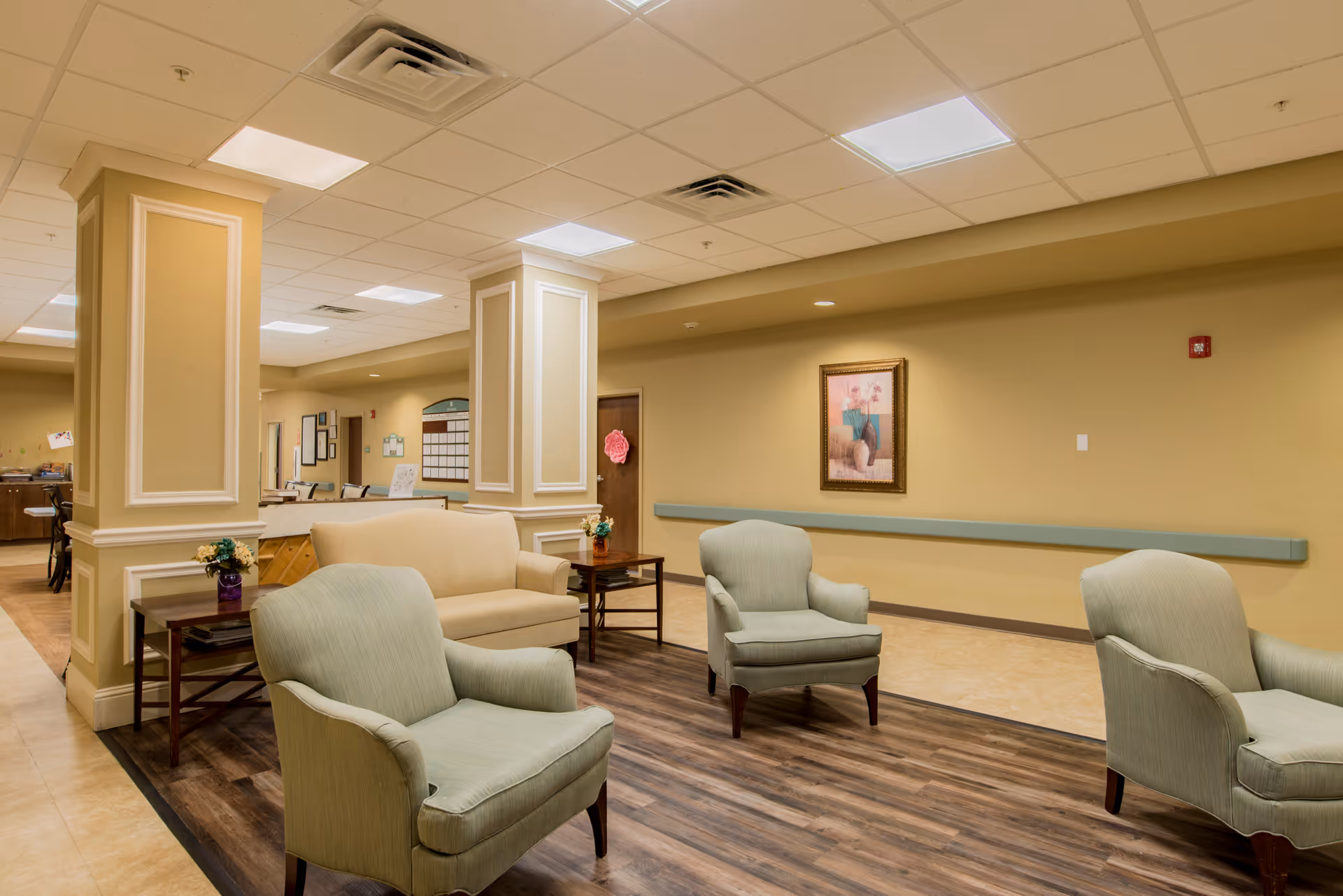 A cozy seating area in a senior living facility with three light green armchairs and a beige loveseat arranged around two wooden side tables with small flower arrangements. The room has beige walls, a wood-patterned floor section, and a framed picture on the wall. The ceiling has recessed lighting and air vents.
