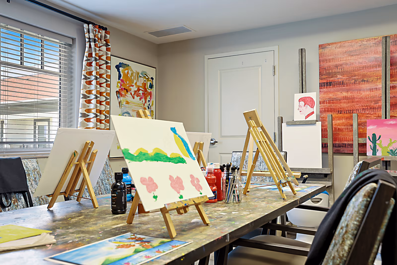 A bright art room with several easels holding canvases, some with paintings in progress. The room has a large window with patterned curtains, colorful artwork on the walls, and a table covered with paint bottles and brushes.