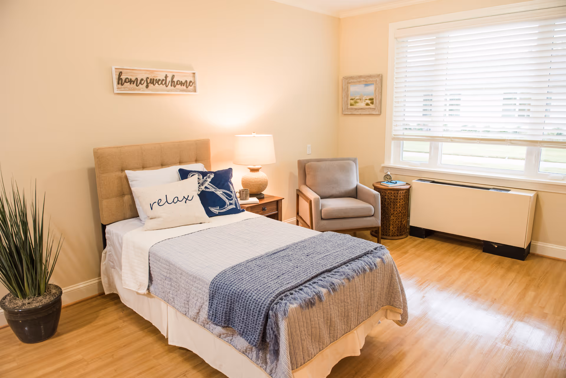 A cozy bedroom with a single bed featuring a beige tufted headboard, white and blue pillows including one with the word 'relax', and a blue throw blanket. Next to the bed is a wooden nightstand with a lamp and a small cup. There is a gray armchair beside a round wooden side table with a clock and a book. A large window with white blinds lets in natural light. The walls are painted light beige with a 'home sweet home' sign above the bed and a framed picture on the adjacent wall. A potted plant is placed on the floor near the bed.