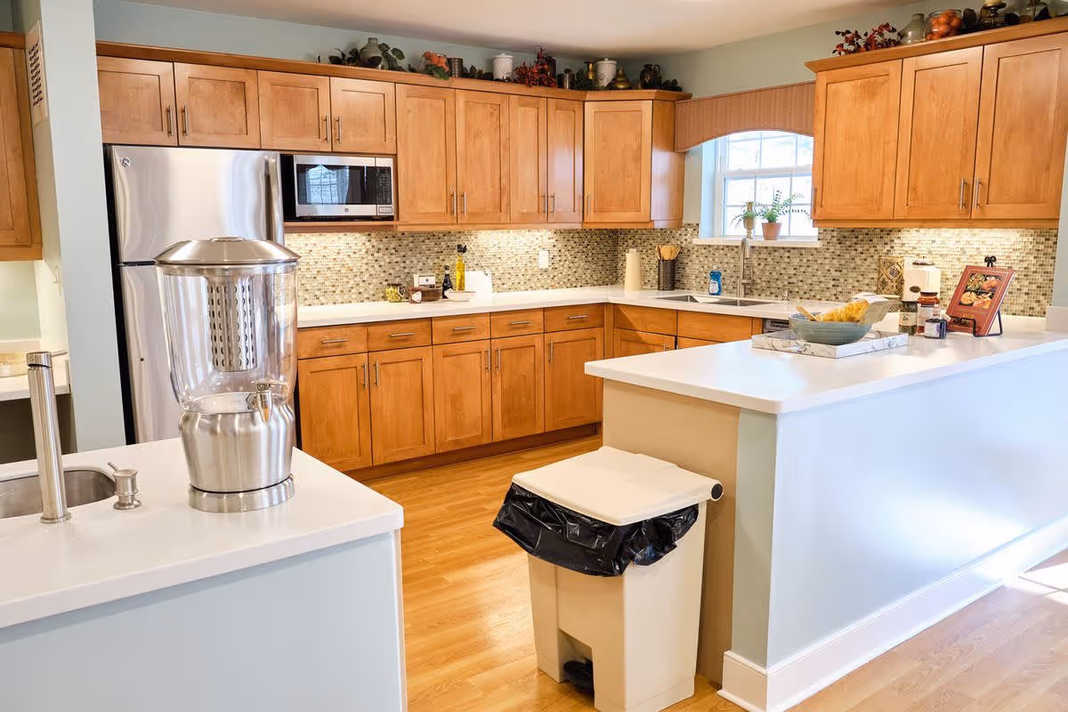 A bright kitchen with wooden cabinets, a mosaic tile backsplash, and white countertops. The kitchen features a stainless steel refrigerator, a microwave, a double sink under a window with potted plants, and various kitchen items on the counters. There is a large water dispenser on the counter in the foreground and a trash bin with a black liner near the counter.
