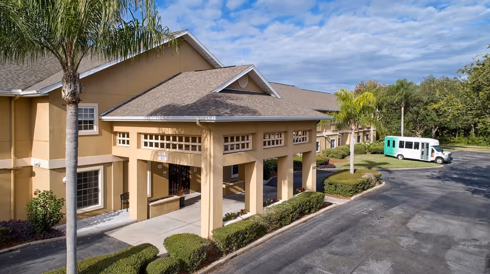 Exterior view of Greenwood Place senior living facility showing a beige building with a covered entrance, palm trees, neatly trimmed bushes, and a parking area with a white and green shuttle bus parked.