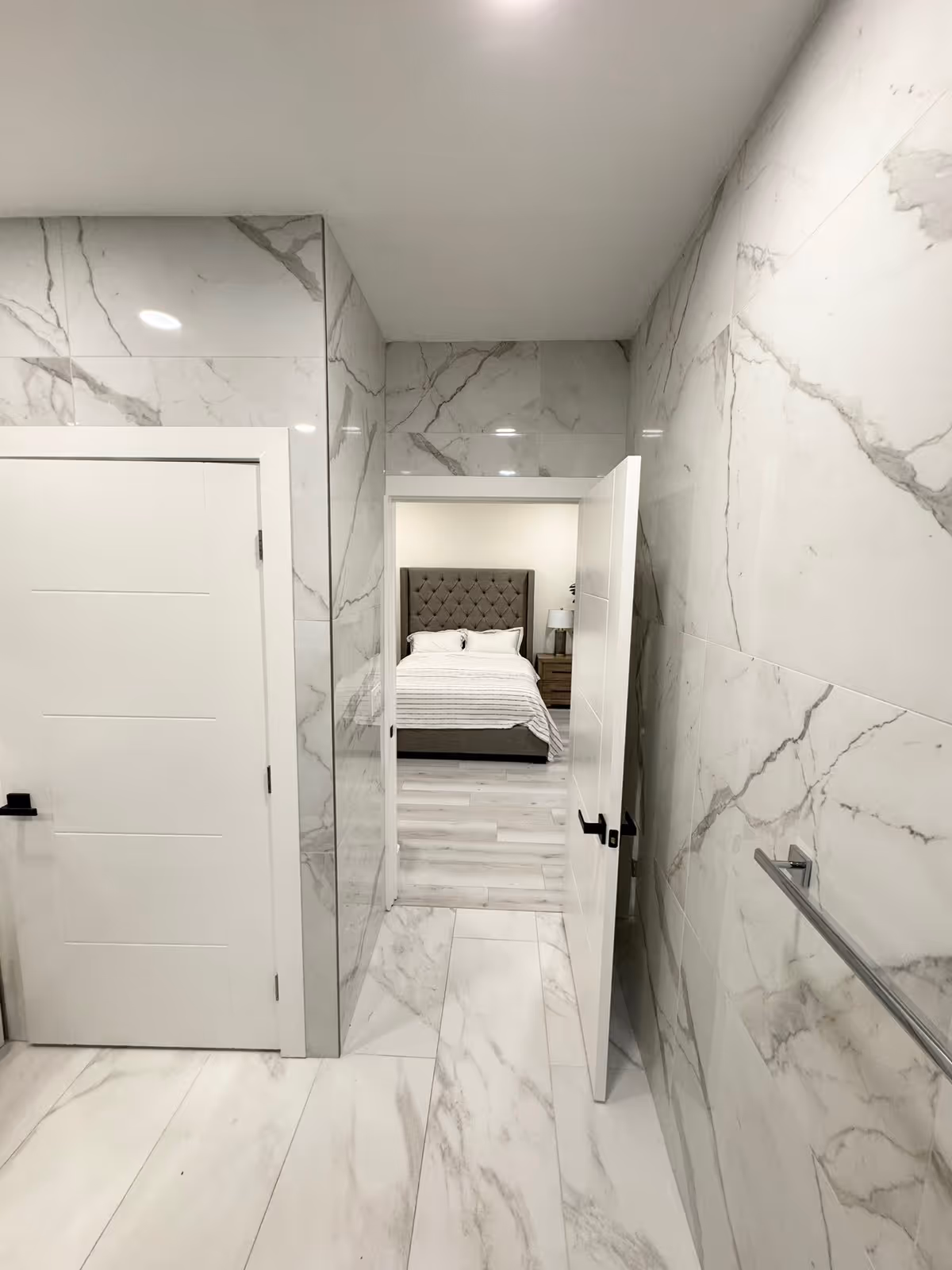 View from a marble-tiled bathroom looking through an open door into a bedroom with a bed featuring a tufted headboard, bedside table, and lamp. The bathroom has white doors and a towel rack on the right wall.