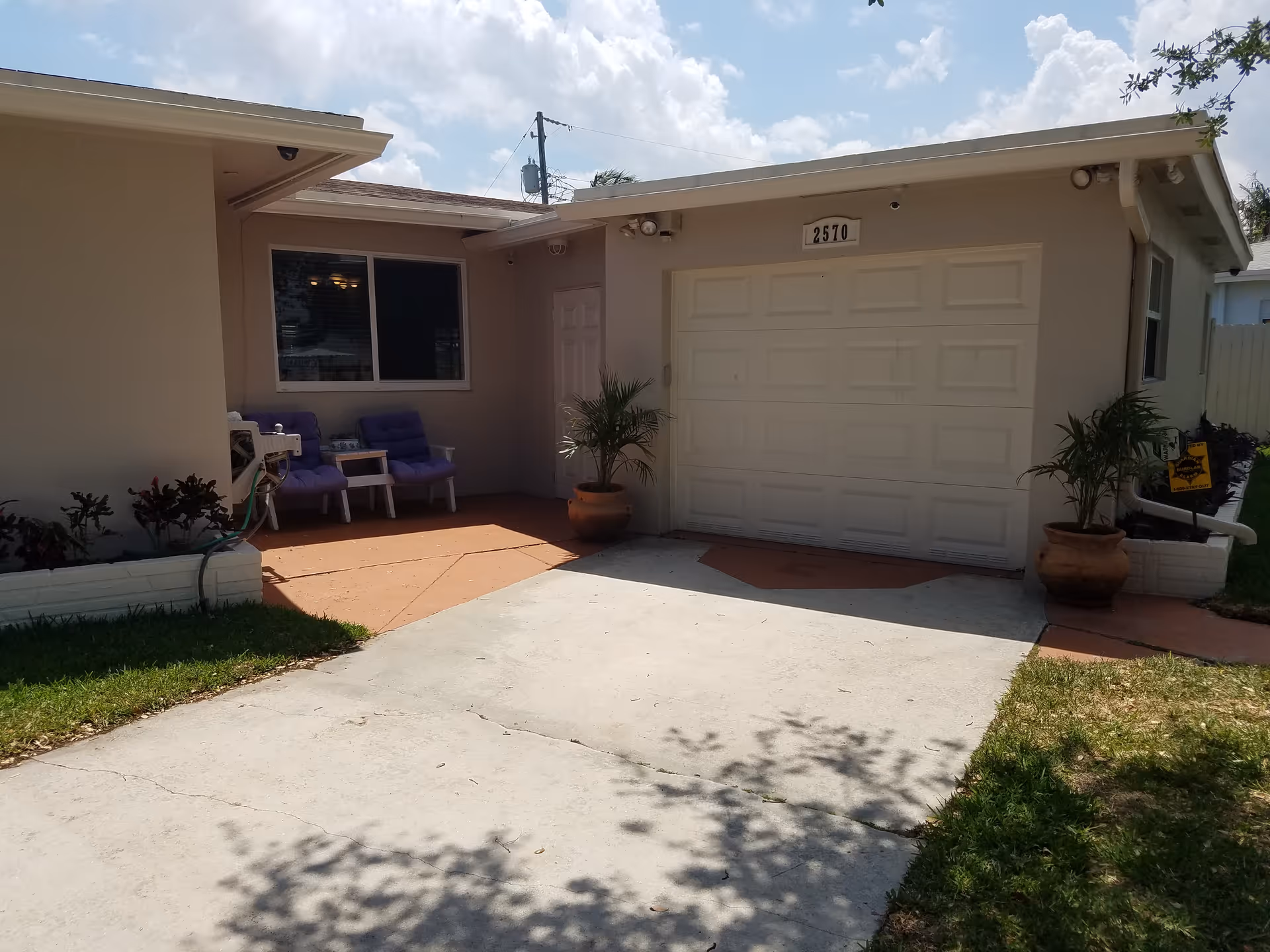 Front exterior of a single-story house with a garage door, driveway, potted plants, and a small seating area on the porch.