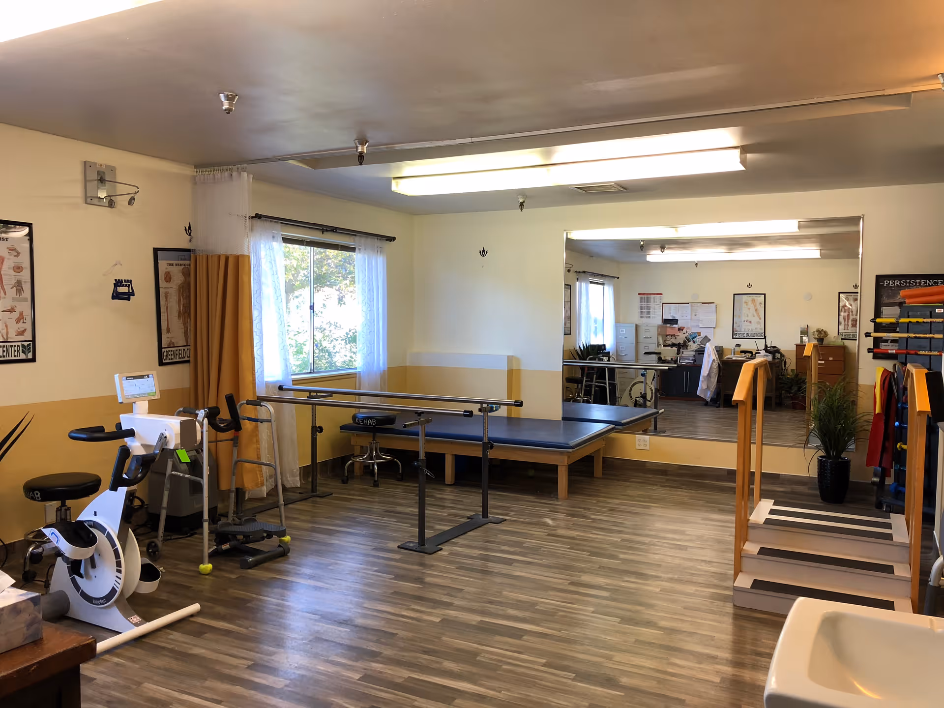 A rehabilitation room with exercise equipment including a stationary bike, parallel bars, a walker, and a step platform. The room has wood-patterned flooring, a large mirror on one wall, and a window with sheer white curtains letting in natural light. There are anatomical posters on the walls and a potted plant near the step platform.