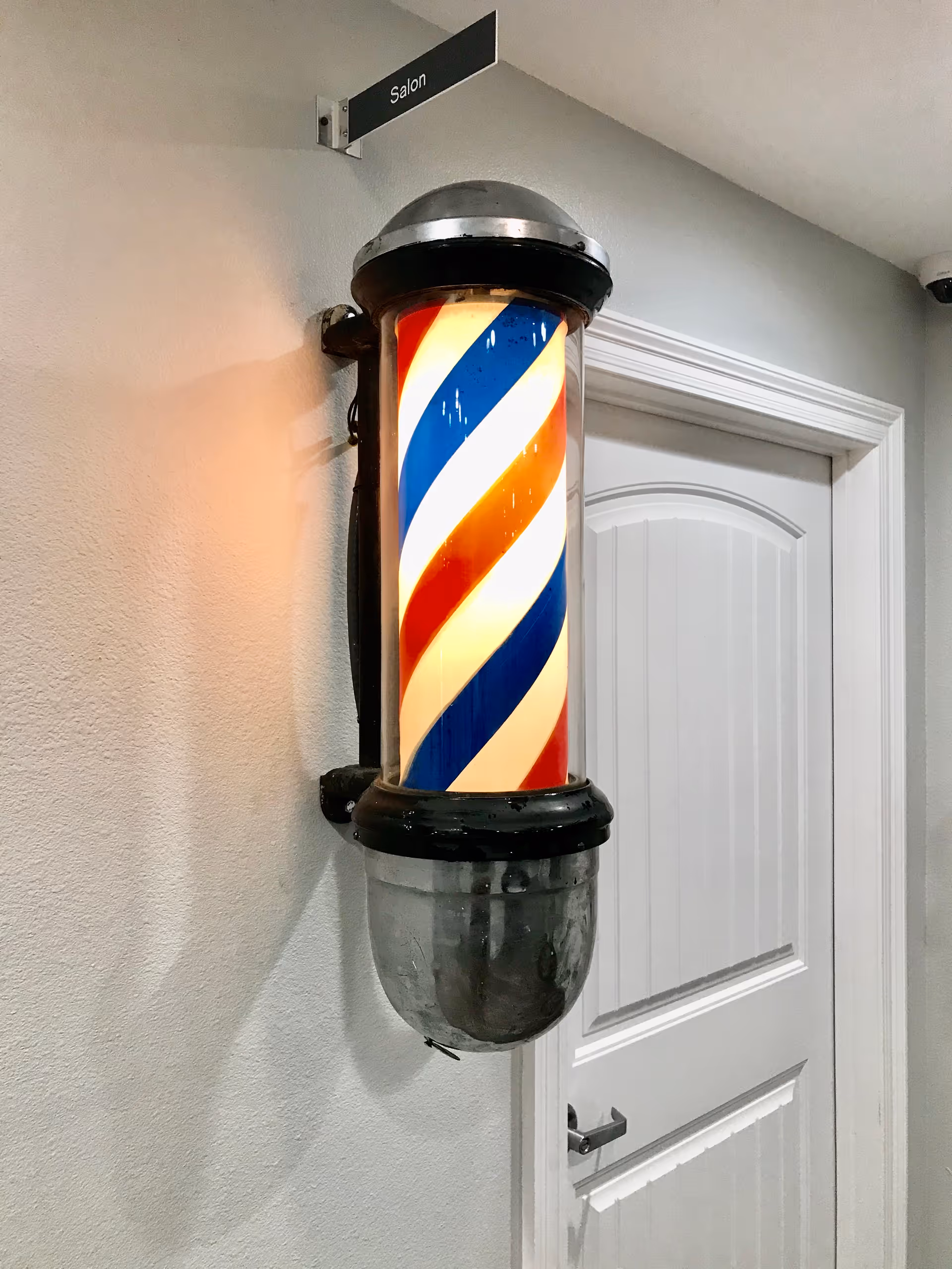 A traditional red, white, and blue barber pole mounted on a wall next to a white door with a sign above it that reads 'Salon'.